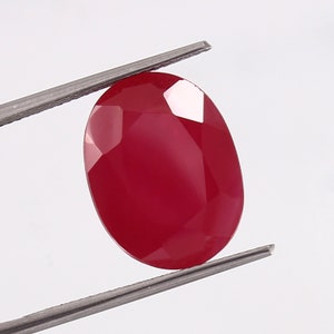Könnte beinhalten: Ein tiefroter ovaler Edelstein mit facettiertem Schliff. Der Edelstein wird in einer Pinzette gehalten.