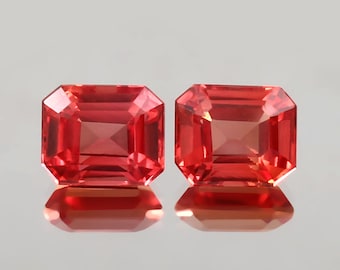 Op de afbeelding: Twee rood-oranje edelstenen met radiant-slijpsel worden getoond tegen een lichte achtergrond. De edelstenen hebben een rechthoekige vorm met afgesneden hoeken en hun facetten reflecteren licht. De stenen liggen naast elkaar, met hun reflecties eronder zichtbaar.