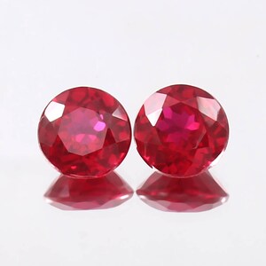 May include: Two round, faceted, deep red rubies are displayed against a white background. The gemstones are highly polished, reflecting light and creating a brilliant sparkle. The rubies are of similar size and shape, showcasing their vibrant colour.
