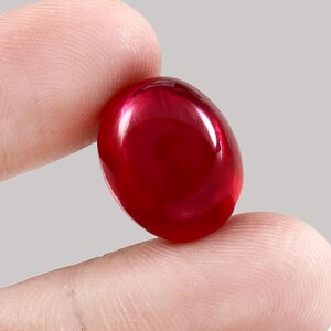 May include: A vibrant, oval-cut, deep red gemstone. The gem is held between two fingers, highlighting its smooth, reflective surface. The stone's colour is a rich, saturated red, with a slight translucence. The gemstone is likely a ruby.