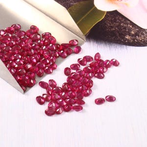 Puede incluir: Un conjunto de rubíes rojos oscuros de corte ovalado. Algunos están en una cuchara plateada, otros dispersos sobre una superficie blanca. Las piedras preciosas facetadas reflejan la luz.