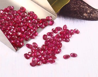 May include: A collection of oval-cut, deep red rubies. Some rubies are in a silver scoop, while others are scattered on a white surface. The gemstones are faceted, reflecting light and showcasing their rich color.