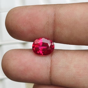 Könnte beinhalten: Ein facettierter, oval geformter, leuchtend roter Edelstein. Der Edelstein wird zwischen zwei Fingern gehalten, was seinen Brillantschliff und seine intensive Farbe zeigt. Die Facetten des Edelsteins reflektieren das Licht und erzeugen einen funkelnden Effekt.
