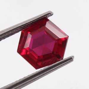 Könnte beinhalten: Ein tief roter Edelstein mit sechseckigem Schliff, gehalten von einer Pinzette.