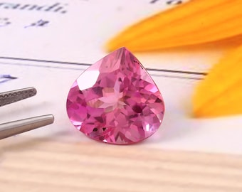 May include: A faceted, teardrop-shaped pink gemstone. The gem is a vibrant shade of pink, with multiple facets catching the light. A pair of tweezers is visible to the left, and a yellow petal is in the background.