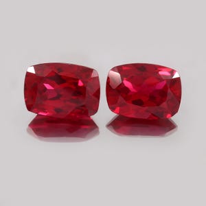 Könnte beinhalten: Zwei facettierte Rubine im Kissenschliff. Die Edelsteine haben eine leuchtend rote Farbe mit sichtbaren inneren Reflexionen. Die Steine liegen auf einer reflektierenden Oberfläche, was ihren Glanz verstärkt. Die Form ist ein modifiziertes Quadrat mit abgerundeten Ecken.