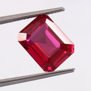 Könnte beinhalten: Ein leuchtend roter Smaragdschliff-Edelstein, der von einer Pinzette gehalten wird. Der Edelstein hat eine tiefe, satte rote Farbe und eine glatte, polierte Oberfläche.
