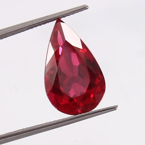 Puede incluir: Una piedra preciosa de rubí en forma de pera con un color rojo intenso. La piedra preciosa está sujeta por unas pinzas.