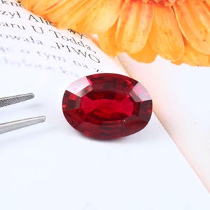 May include: A faceted, oval-shaped, deep red gemstone. The ruby is set against a white background with a flower and text in the background. A pair of tweezers is in the lower left corner.