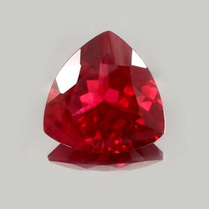Könnte beinhalten: Ein facettierter, dreieckiger Rubin-Edelstein. Der Edelstein hat eine tiefe, lebendige rote Farbe mit mehreren reflektierenden Oberflächen. Der Stein liegt vor einem hellgrauen Hintergrund, wobei sich sein Spiegelbild darunter zeigt.
