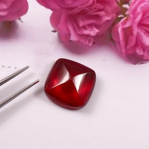 May include: A square, deep red gemstone with a polished surface. The gem is held by tweezers, with pink roses in the background. The gem's facets reflect light, creating a brilliant shine. This gemstone is likely for jewelry making.