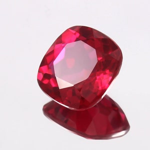 Könnte beinhalten: Ein facettierter Rubin-Edelstein im Kissenschliff. Der Edelstein hat eine leuchtend rote Farbe mit mehreren Facetten, die das Licht reflektieren. Der Rubin liegt auf einer reflektierenden Oberfläche, wodurch ein Spiegelbild des Steins entsteht.