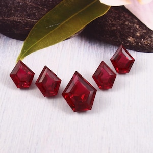 Könnte beinhalten: Fünf facettierte, tiefrote Edelsteine, die in einer Reihe angeordnet sind. Jeder Edelstein hat eine einzigartige rautenförmige Form mit mehreren Facetten, die das Licht reflektieren. Die Edelsteine liegen auf einer weißen und hellblauen Oberfläche, mit einem grünen Blatt und einer Blume im Hintergrund.