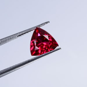 Könnte beinhalten: Ein tiefroter dreieckiger Edelstein, der von einer Pinzette vor einem weißen Hintergrund gehalten wird.