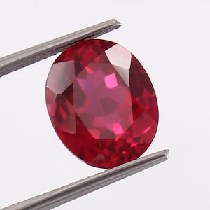 Könnte beinhalten: Ein facettierter, oval geschliffener Rubin-Edelstein, der von einer Metallpinzette gehalten wird. Der Rubin hat eine tiefe, gesättigte rote Farbe mit mehreren Facetten, die das Licht reflektieren. Die Pinzette ist silberfarben.