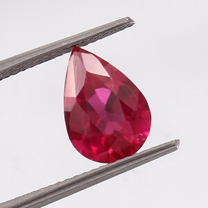 Könnte beinhalten: Ein birnenförmiger Rubin-Edelstein mit einer tief roten Farbe. Der Edelstein wird von einer Pinzette gehalten.