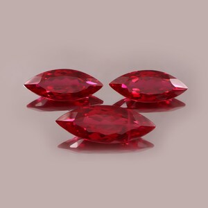 Könnte beinhalten: Drei facettierte Edelsteine im Marquise-Schliff in einem leuchtenden Rotton. Die Edelsteine sind in einer dreieckigen Anordnung angeordnet, wobei jeder das Licht reflektiert und einen Schatten auf die darunter liegende Oberfläche wirft. Der Hintergrund ist eine weiche, neutrale Farbe.