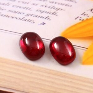 Puede incluir: Dos gemas ovaladas de color rojo intenso con un acabado brillante. Las gemas se colocan sobre una página de libro con texto italiano y un pétalo amarillo.