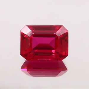 May include: A vibrant red emerald-cut gemstone with a high polish. The gemstone is set against a white background.