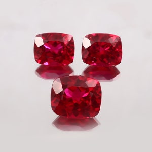 Könnte beinhalten: Drei rote Kissen-Schliff-Edelsteine auf weißem Hintergrund. Die Edelsteine sind facettiert und haben einen hohen Glanz.