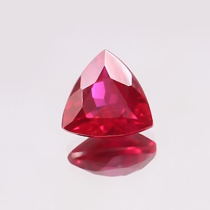 Könnte beinhalten: Ein leuchtend roter, dreieckiger Edelstein mit facettiertem Schliff. Der Edelstein ist vor einem weißen Hintergrund platziert.