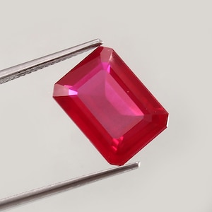 Könnte beinhalten: Ein leuchtend roter Edelstein mit einem rechteckigen Smaragdschliff. Der Edelstein wird in einer Pinzette vor einem weißen Hintergrund gehalten.