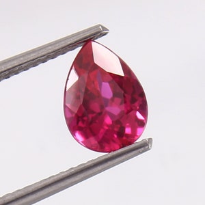 Puede incluir: Una piedra preciosa de rubí en forma de pera con un color rojo intenso. La piedra preciosa está sujeta por unas pinzas.