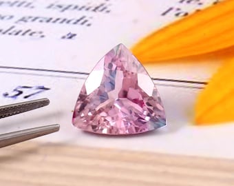 May include: A triangular, faceted pink gemstone. The gem is set against a backdrop of a book page and a yellow flower petal. The gem is held by a pair of silver tweezers.