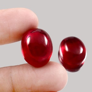 Puede incluir: Dos gemas ovaladas de color rojo intenso con un acabado pulido y brillante. Las gemas se muestran sobre un fondo neutro, resaltando su rico color y superficie lisa. Una gema se sostiene entre los dedos.
