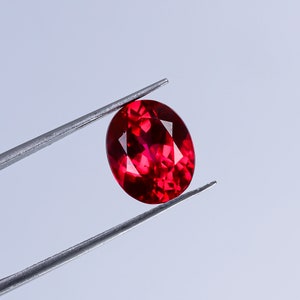 Könnte beinhalten: Ein leuchtend roter ovaler Edelstein, der von einer Pinzette gehalten wird. Der Edelstein ist facettiert und reflektiert Licht.