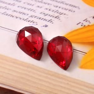 May include: Two faceted, teardrop-shaped, deep red gemstones. The gems have a brilliant cut, reflecting light. They are set against a backdrop of a book and a yellow flower petal, creating a striking contrast.