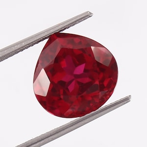 Könnte beinhalten: Ein tief roter birnenförmiger Edelstein mit facettiertem Schliff. Der Edelstein wird in einer Pinzette gehalten.