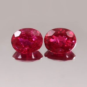 Puede incluir: Dos rubíes ovalados y facetados. Las piedras preciosas son de un rojo intenso y vibrante con reflejos internos, sobre una superficie reflectante. Las gemas se muestran una al lado de la otra, mostrando su claridad y color.