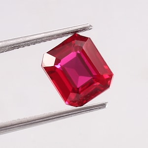 Könnte beinhalten: Ein leuchtend roter Smaragdschliff-Edelstein, der von einer Pinzette gehalten wird. Der Edelstein hat eine tiefe, satte rote Farbe und eine hohe Klarheit.