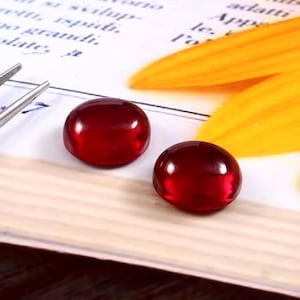 May include: Two polished, round, deep red gemstones. The gems are smooth and reflective, with a slight dome shape. They sit on a white surface with text and a yellow petal in the background. A pair of tweezers is visible on the left.