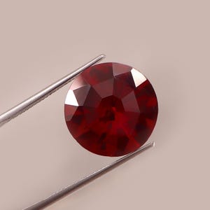 Könnte beinhalten: Ein runder, facettierter, tiefroter Edelstein, der von einer silberfarbenen Metallpinzette gehalten wird. Der Stein weist einen Brillantschliff mit zahlreichen dreieckigen und rautenförmigen Facetten auf, die das Licht reflektieren.