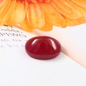 Può includere: Una gemma liscia, di forma ovale, rosso scuro. La pietra lucida riflette la luce, su una superficie bianca con un fiore arancione sfocato sullo sfondo. La gemma è probabilmente per la creazione di gioielli.
