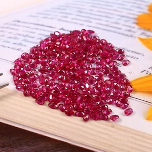 Könnte beinhalten: Ein Haufen kleiner, oval geformter, tiefroter Edelsteine. Die Edelsteine sind auf einer hellen Oberfläche, möglicherweise einer Buchseite, verstreut, mit einem unscharfen Hintergrund. Bei den Edelsteinen handelt es sich wahrscheinlich um Rubine, die für ihre leuchtende Farbe und Verwendung in Schmuck bekannt sind.