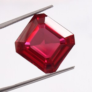 Könnte beinhalten: Ein facettierter, quadratisch geschliffener Rubin, gehalten von einer Pinzette. Der Edelstein hat eine tiefe, gesättigte rote Farbe mit scharfen Kanten und einer reflektierenden Oberfläche. Der Rubin wird vor einem weißen Hintergrund präsentiert.
