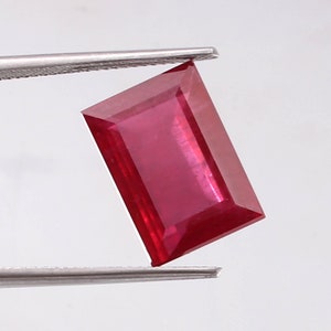 May include: A vibrant red rectangular gemstone with a faceted cut. The gemstone is held by tweezers against a white background.