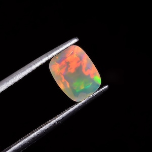 Könnte beinhalten: Ein weißer Opal-Edelstein mit einem Regenbogen aus Farben, darunter Orange, Grün und Blau, wird von einer Pinzette vor einem schwarzen Hintergrund gehalten.