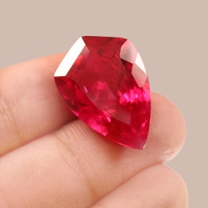Könnte beinhalten: Ein tief roter Edelstein mit einem facettierten Schliff, der die Form einer Träne hat. Der Edelstein wird in einer Hand gehalten.