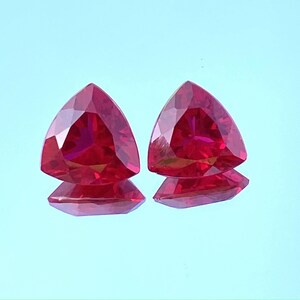 Könnte beinhalten: Zwei dreieckige rote Edelsteine mit Facettenschliff. Die Edelsteine sind vor einem hellblauen Hintergrund platziert.
