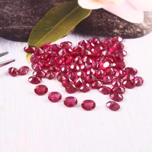 May include: A collection of oval-cut, deep red rubies. The gemstones are scattered across a white surface, with a few visible near a pair of tweezers. The rubies are faceted, reflecting light, and are a rich, vibrant color.