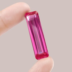 May include: A vibrant pink gemstone with a rectangular cut and a faceted finish. The gemstone is held between two fingers.