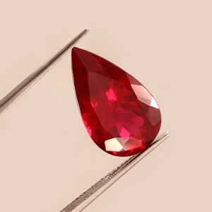 May include: A pear-shaped ruby gemstone with a deep red colour. The gemstone is held in place by a pair of tweezers.