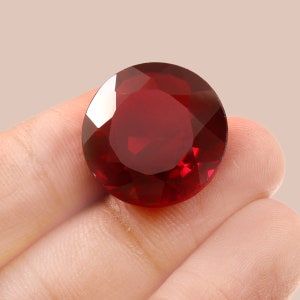 May include: A deep red gemstone with a round, faceted cut. The gemstone is held in a hand.
