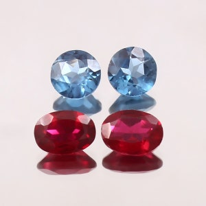 Può includere: Quattro pietre preziose: due gemme rotonde blu chiaro e due gemme rosse ovali. Le gemme sono esposte su una superficie riflettente, creando riflessi speculari di ogni pietra. La luce cattura le sfaccettature delle gemme.