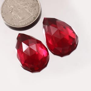 Könnte beinhalten: Zwei facettierte, tropfenförmige, tiefrote Edelsteine. Die Edelsteine liegen neben einer Vierteldollarmünze zum Größenvergleich. Die Edelsteine haben eine Hochglanzoberfläche und reflektieren das Licht, wodurch ihre komplizierten Schnitte zur Geltung kommen. Ideal für Schmuckherstellung.