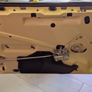 May include: Interior car door panel in a light yellow color. The door panel has various holes and mounting points for hardware. A metal latch mechanism and a black protective cover are visible.